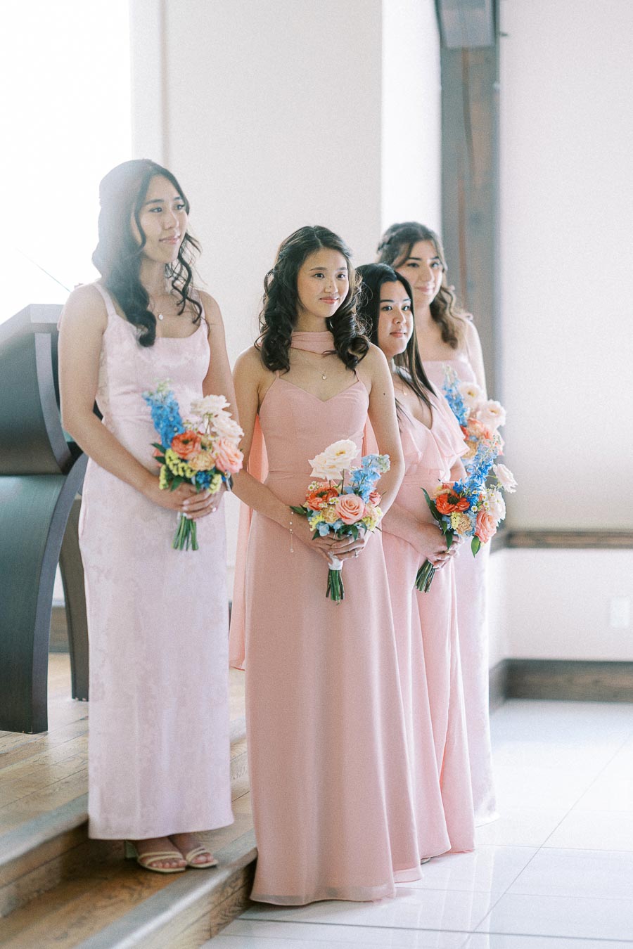Four bridesmaids in blush pink dresses hold colorful bouquets while standing on indoor steps.