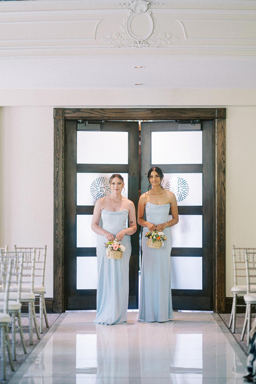 Two bridesmaids in pale blue gowns stand at chapel doors holding flower baskets before the aisle