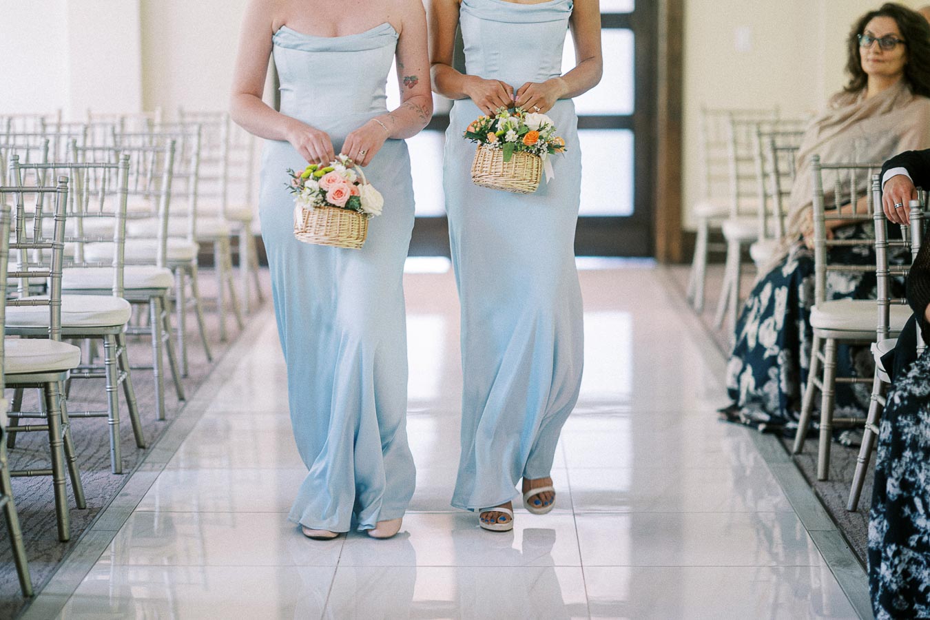 Two bridesmaids in pale blue dresses carry flower baskets down the aisle between silver chairs.