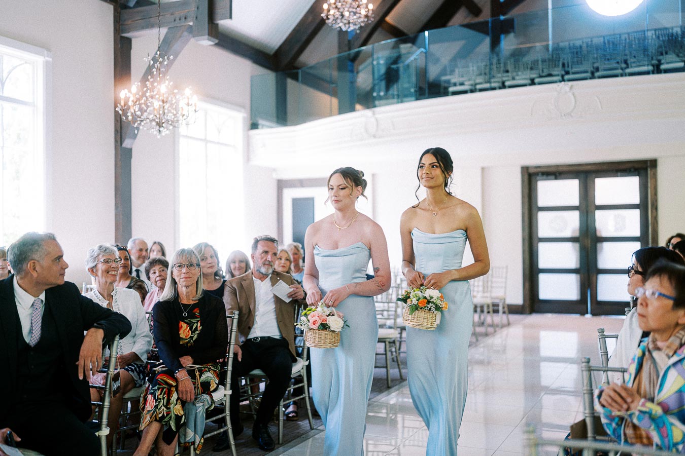 Two bridesmaids in pale blue dresses carry flower baskets down the aisle as seated guests watch.