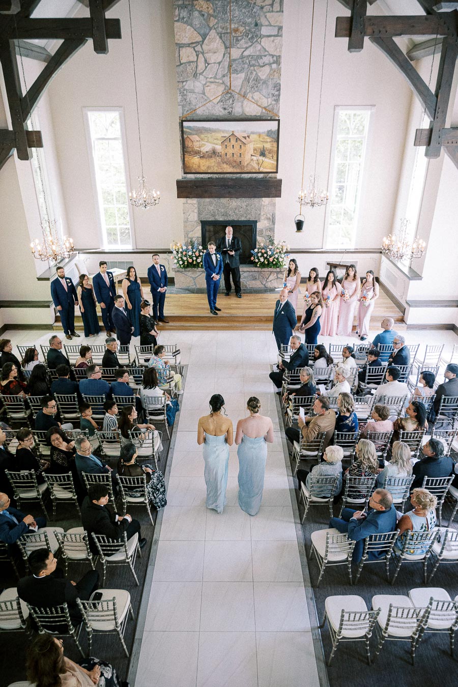 Two bridesmaids walk down the aisle during an indoor chapel ceremony with guests and wedding party watching.