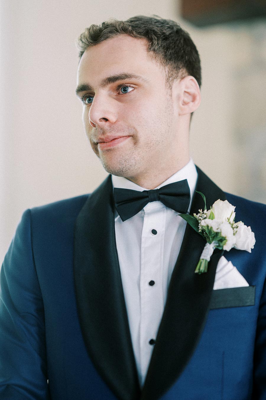 Groom in a blue tuxedo with black bow tie and white rose boutonniere looking to the side