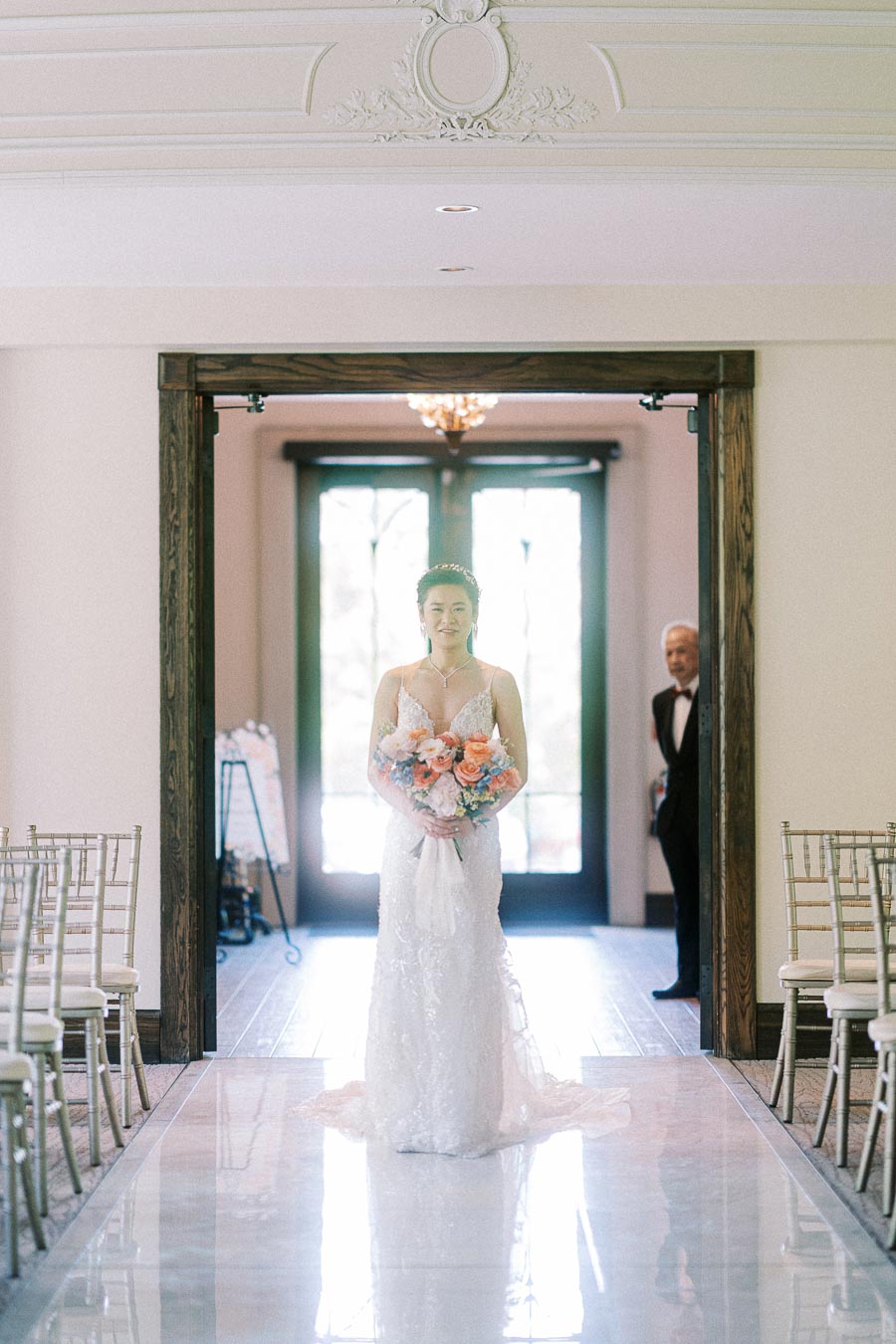 Bride holding a pastel bouquet at the start of the ceremony aisle as an older man waits behind her.