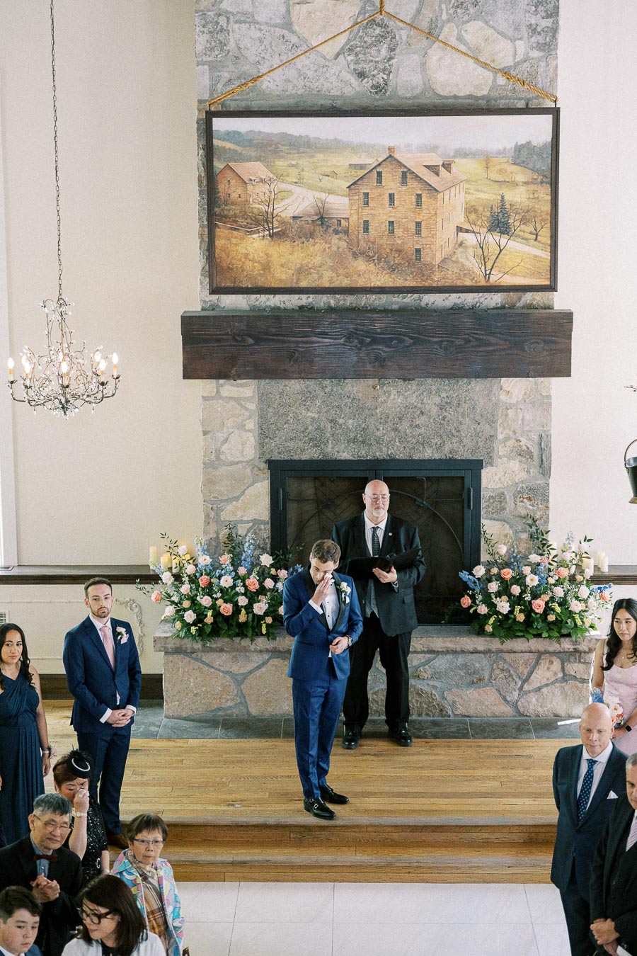 Groom in a blue suit wipes his face at the altar beside an officiant, with guests seated in a stone chapel room.