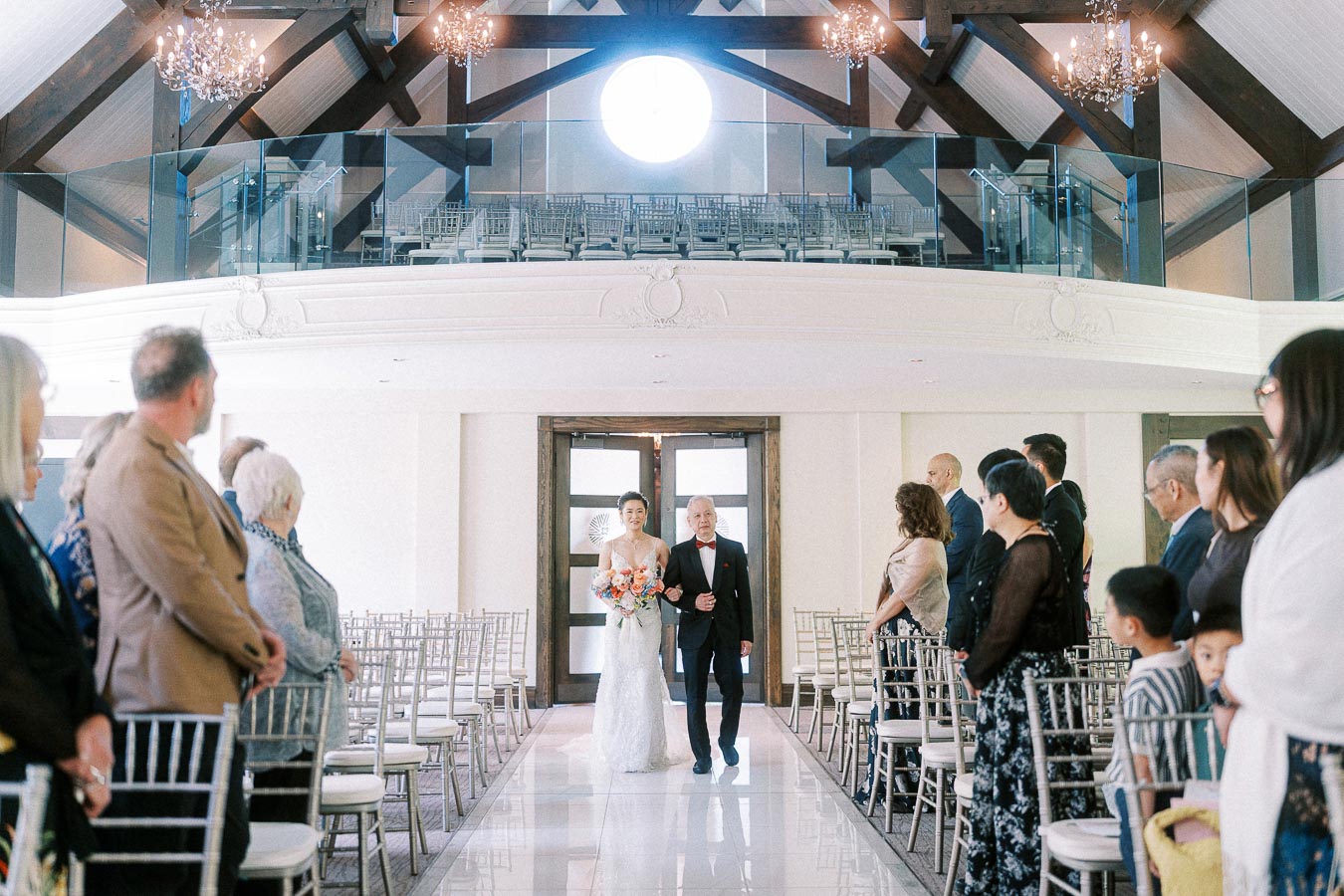 Bride walks down the chapel aisle with an older man as guests stand beside silver chairs under chandeliers.