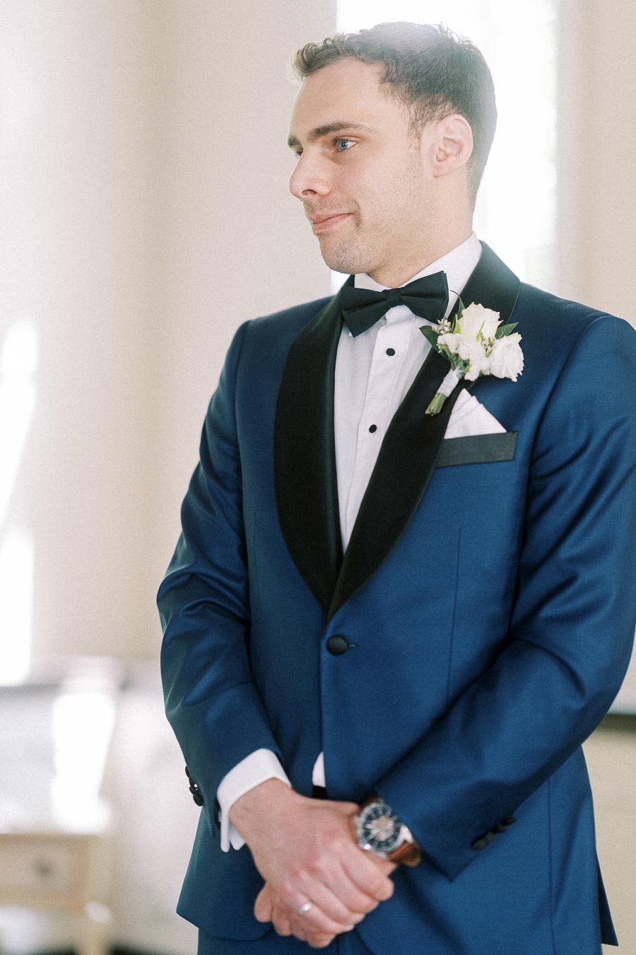 Groom in a blue tuxedo with black bow tie, white boutonniere, folded pocket square, and wristwatch indoors