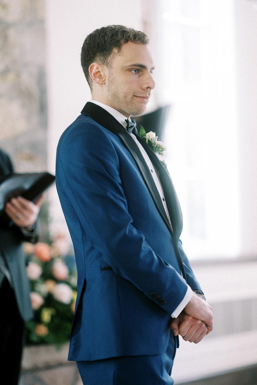 Groom in a blue tuxedo stands at the altar during an indoor wedding ceremony.