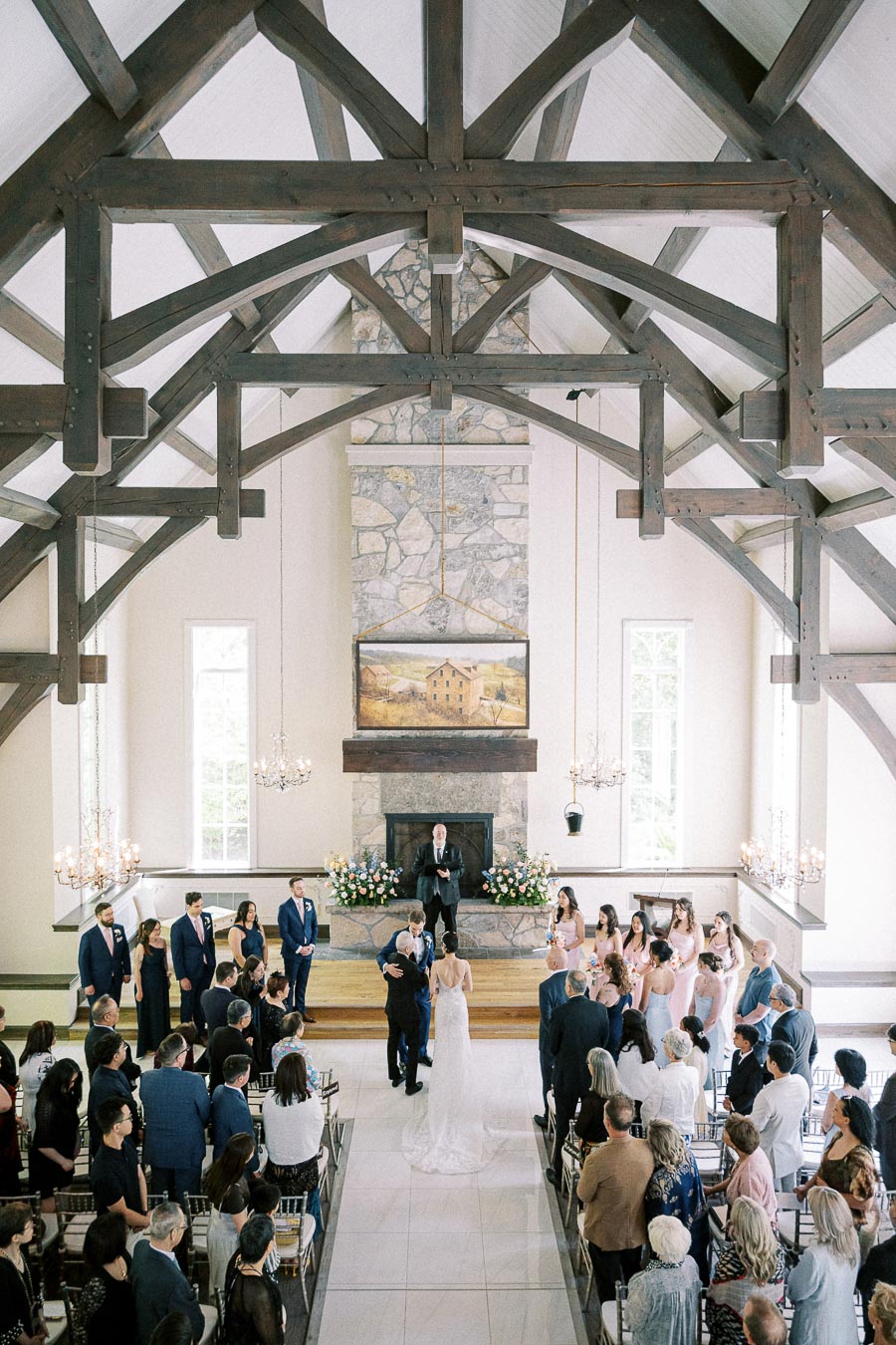 Wide chapel view as a bride walks down the aisle with a companion beneath timber beams and chandeliers