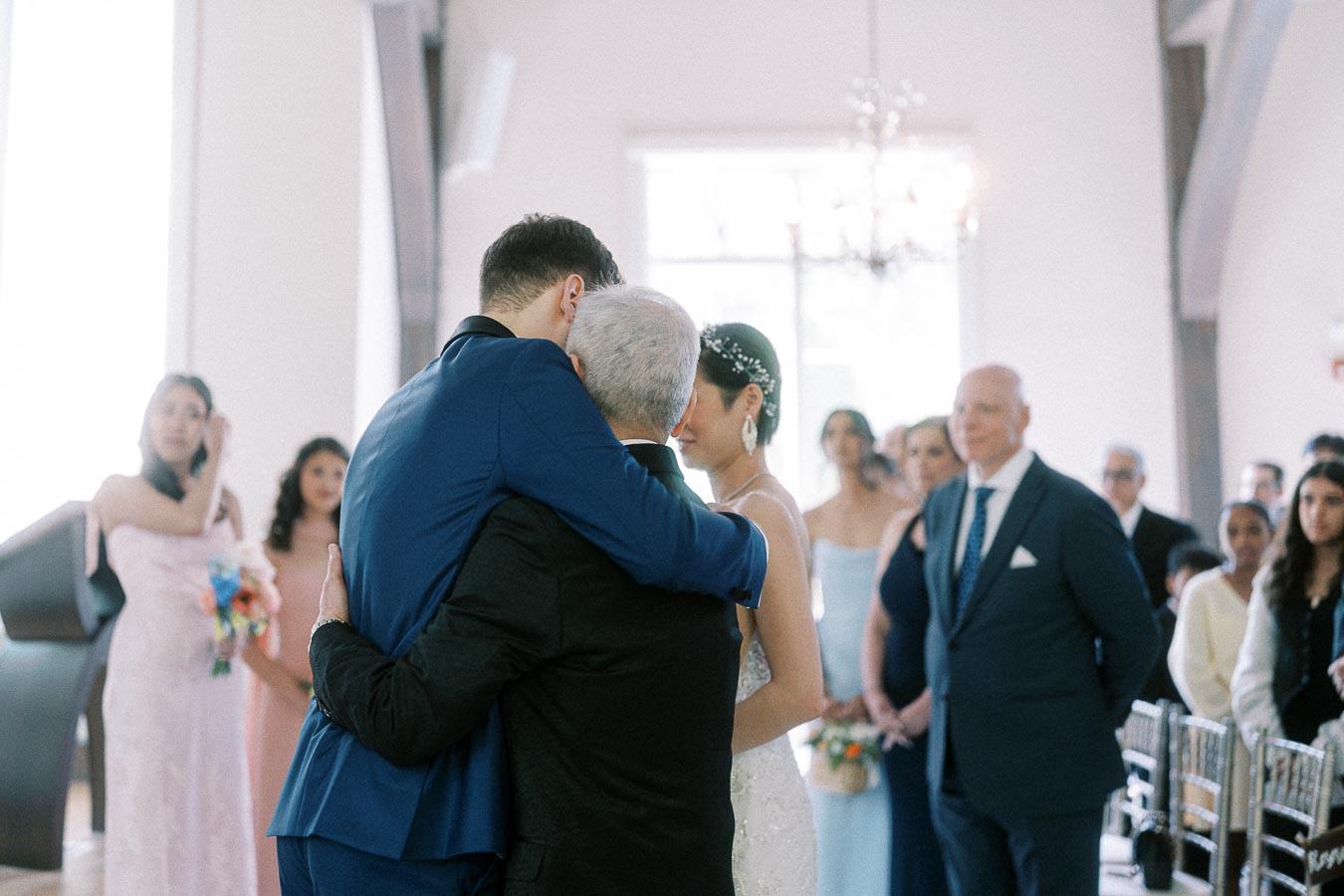 Groom in a blue suit hugs an older man beside the bride during an indoor wedding ceremony.
