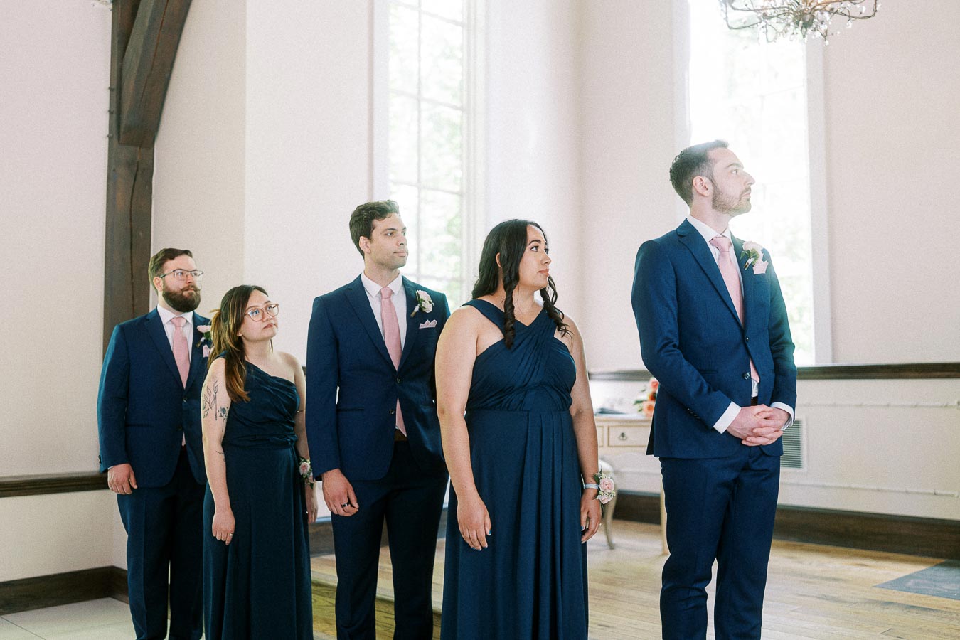 Wedding party in navy attire stands inside a bright room with tall windows and wood beams.