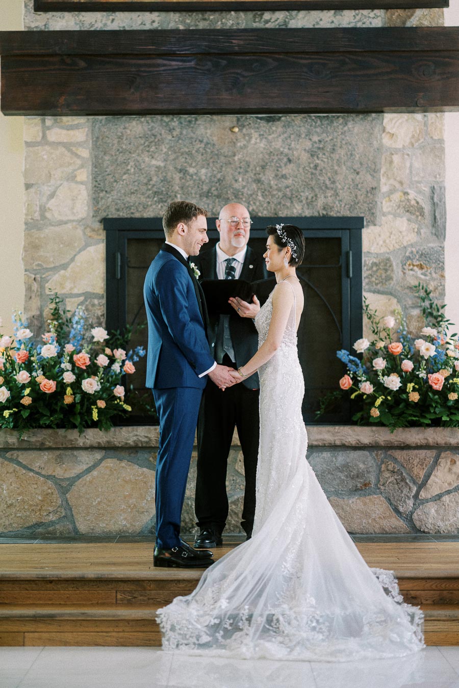 Miffy and Rawle hold hands during their indoor wedding ceremony before a stone fireplace and floral arrangements.