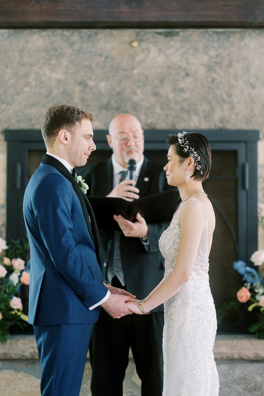 Miffy and Rawle hold hands during their indoor wedding ceremony with an officiant standing behind them.