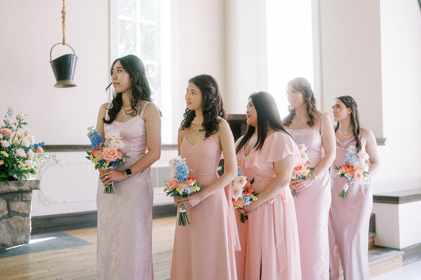 Five bridesmaids in blush pink dresses hold colorful bouquets inside a bright chapel at Ancaster Mill.