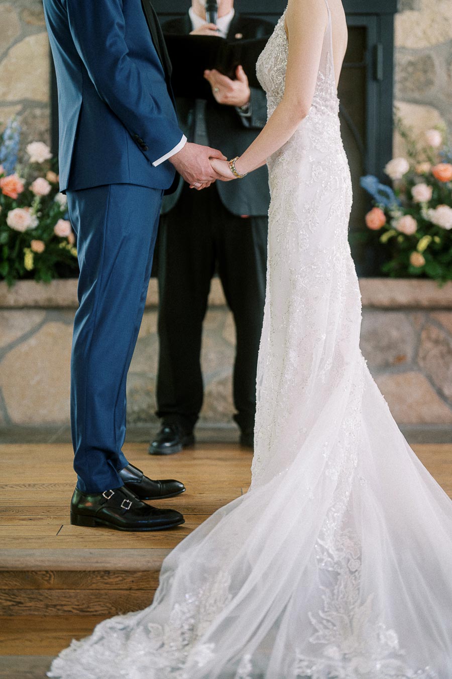 Bride and groom hold hands during their indoor ceremony beside an officiant, with floral arrangements and a stone fireplace.