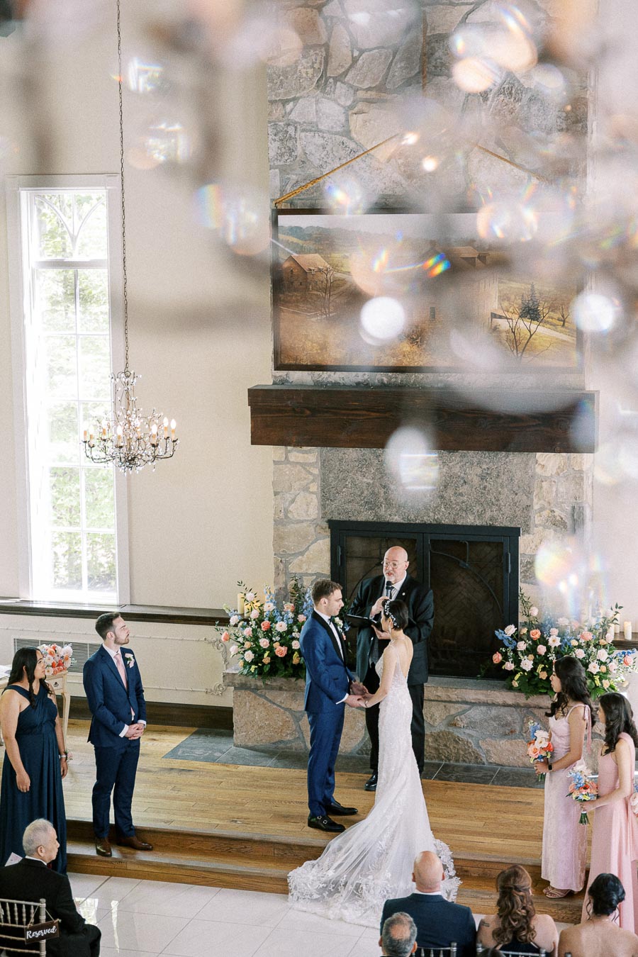 Miffy and Rawle hold hands during their indoor wedding ceremony before a stone fireplace at Ancaster Mill.