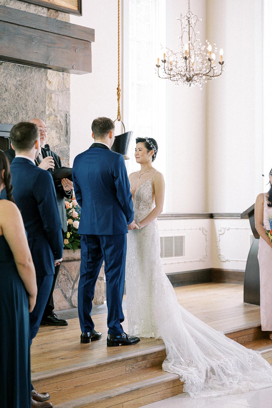 Bride and groom stand before an officiant during an indoor wedding ceremony with chandelier and stone fireplace.