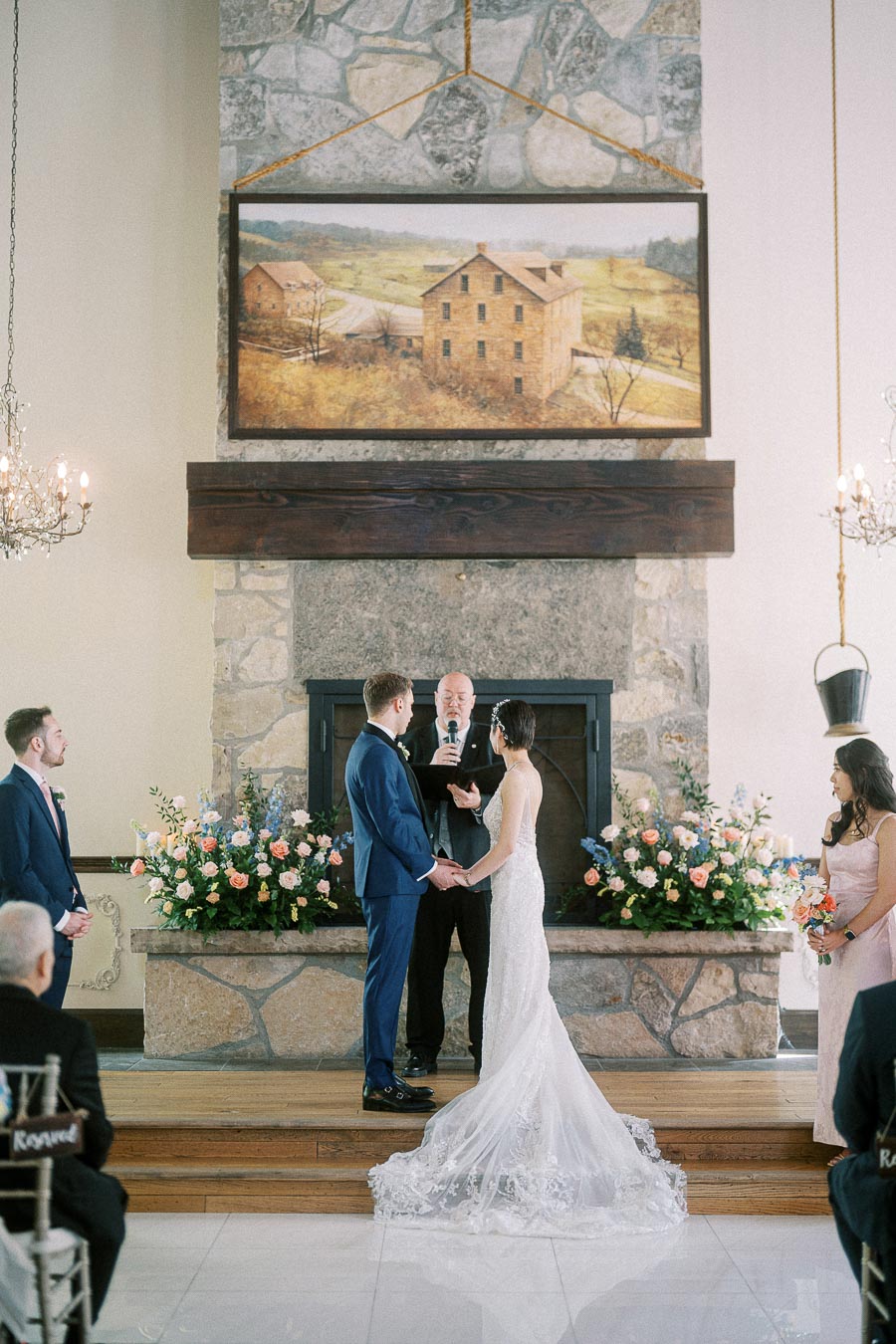 Bride and groom hold hands during their indoor ceremony before a stone fireplace with floral arrangements.