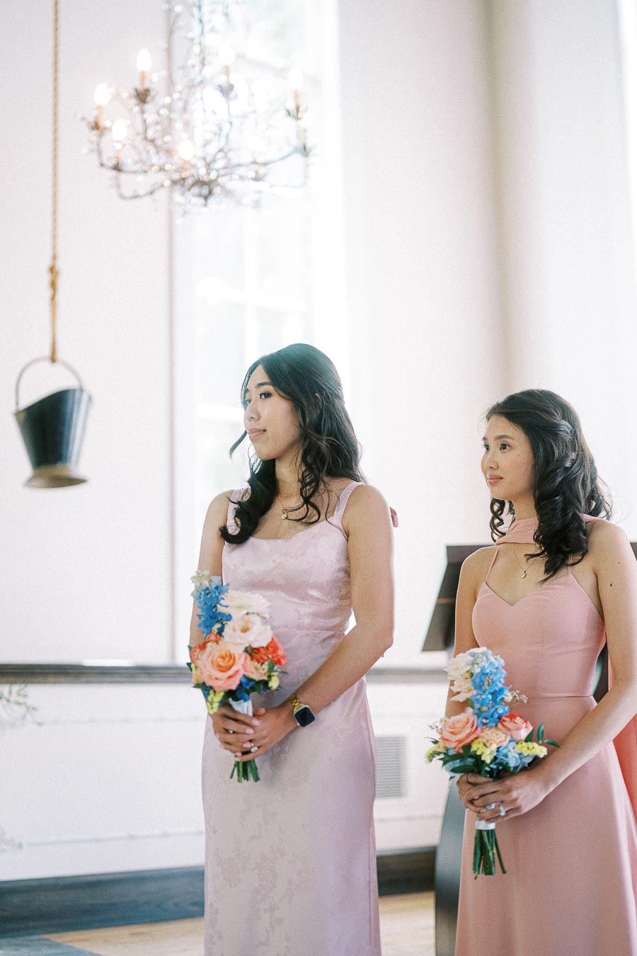 Two bridesmaids in pink dresses hold colorful bouquets during an indoor wedding ceremony