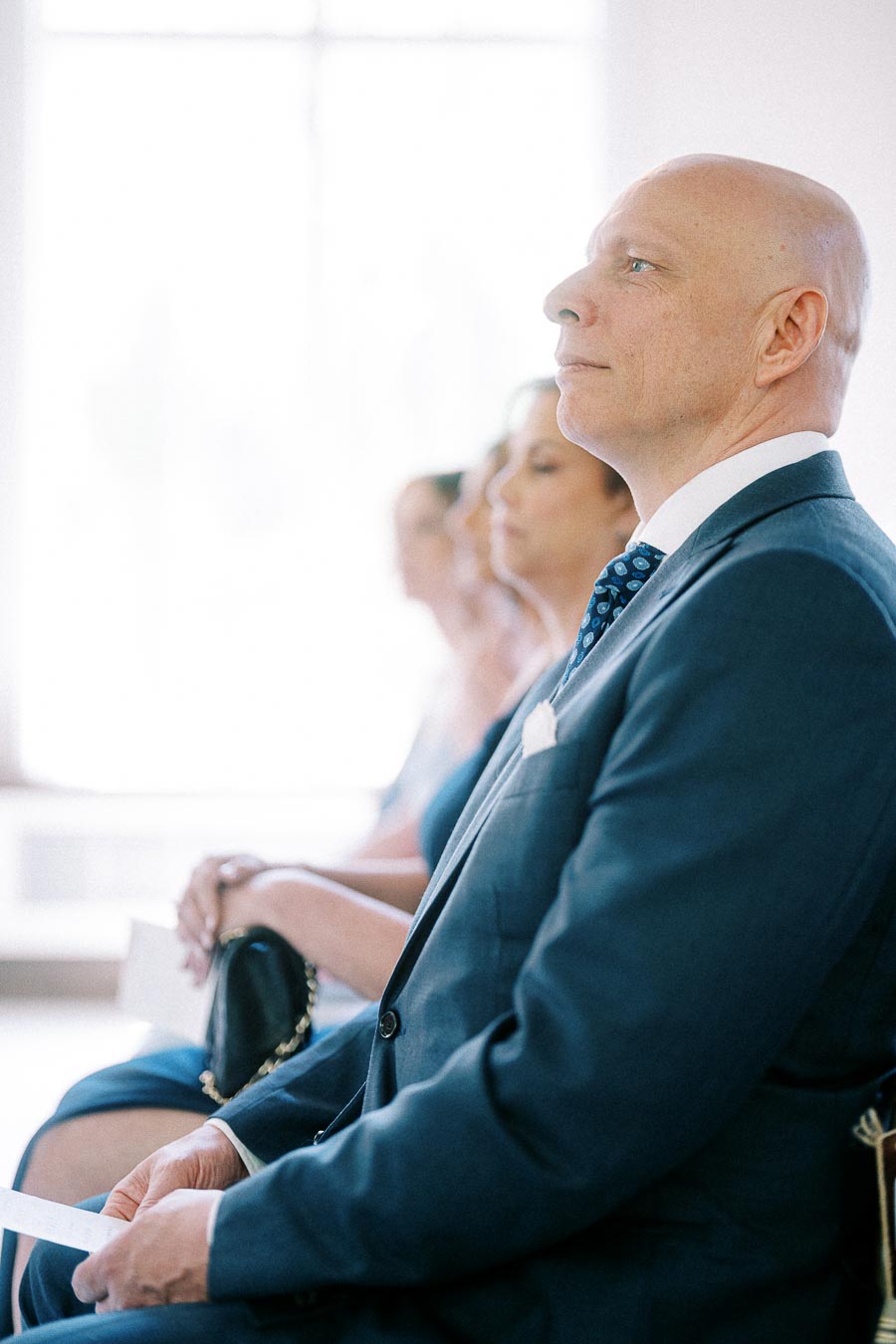 Bald man in a navy suit sits beside guests during a bright indoor wedding ceremony.