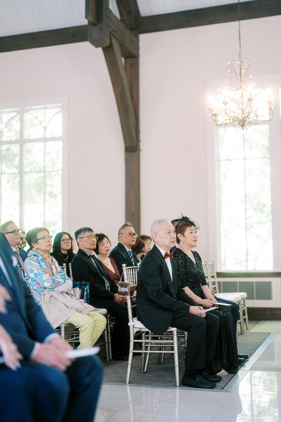 Wedding guests sit in white chairs beneath timber beams and a chandelier during an indoor ceremony.
