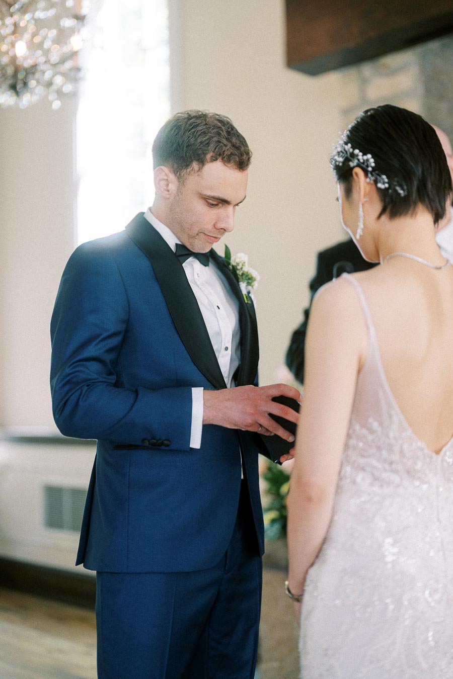 Groom in a blue tuxedo places a wedding ring on the bride’s finger during an indoor ceremony.