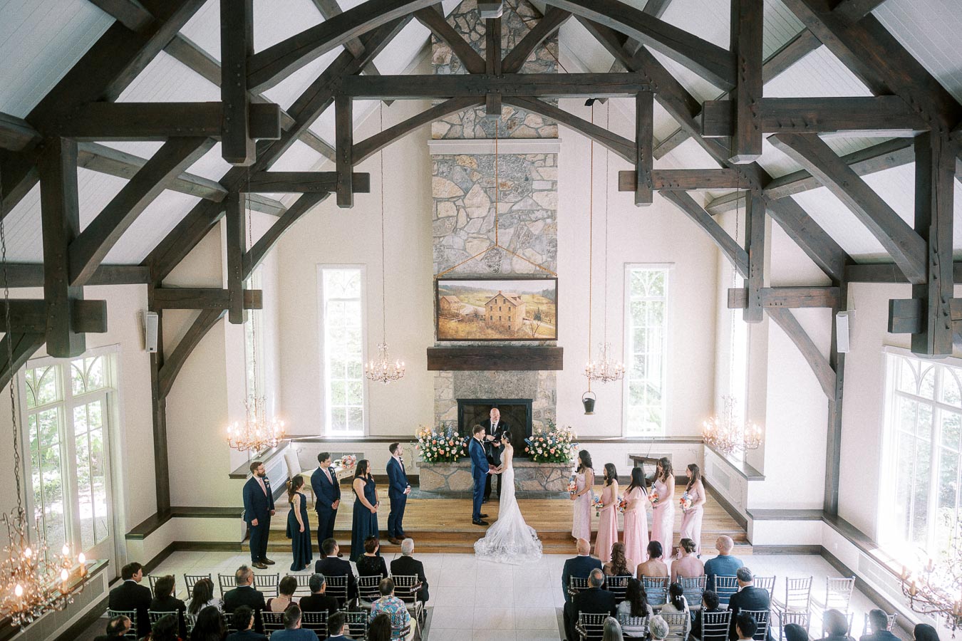 Bride and groom exchange vows in a timber-framed chapel with wedding party and guests seated below.