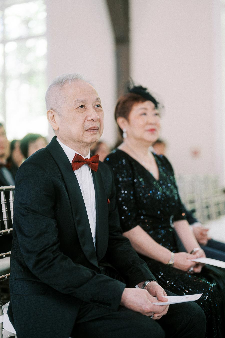 Older wedding guests seated during the ceremony, holding programs and facing forward in a bright chapel space.