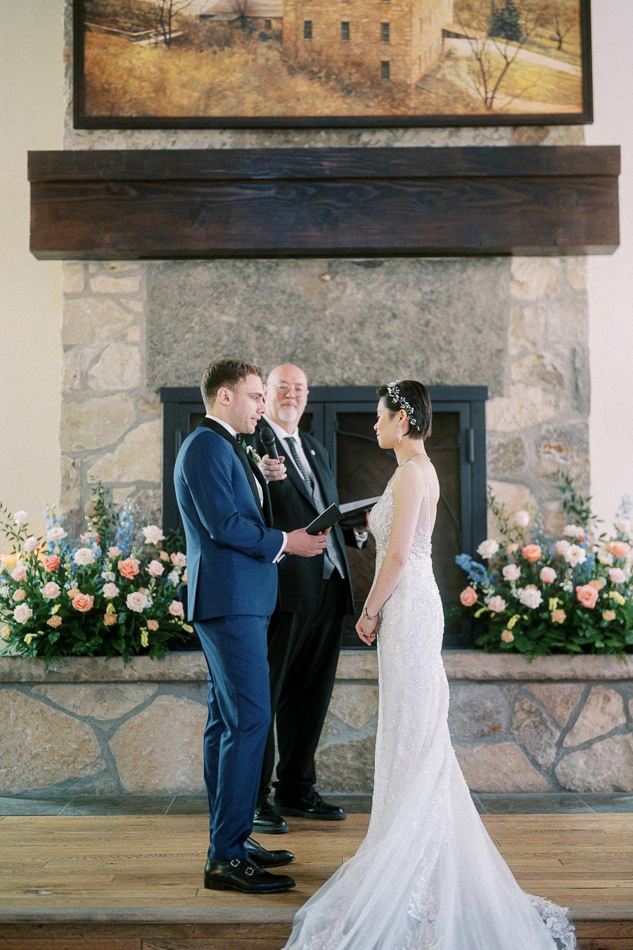 Bride and groom exchange vows before an officiant by a stone fireplace with pastel flowers.