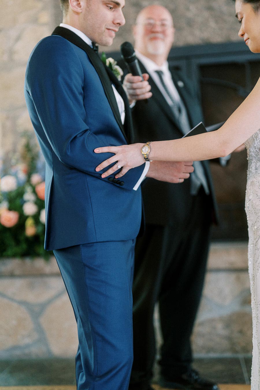 Bride places her hand on the groom’s blue suit during an indoor wedding ceremony with an officiant holding a microphone.