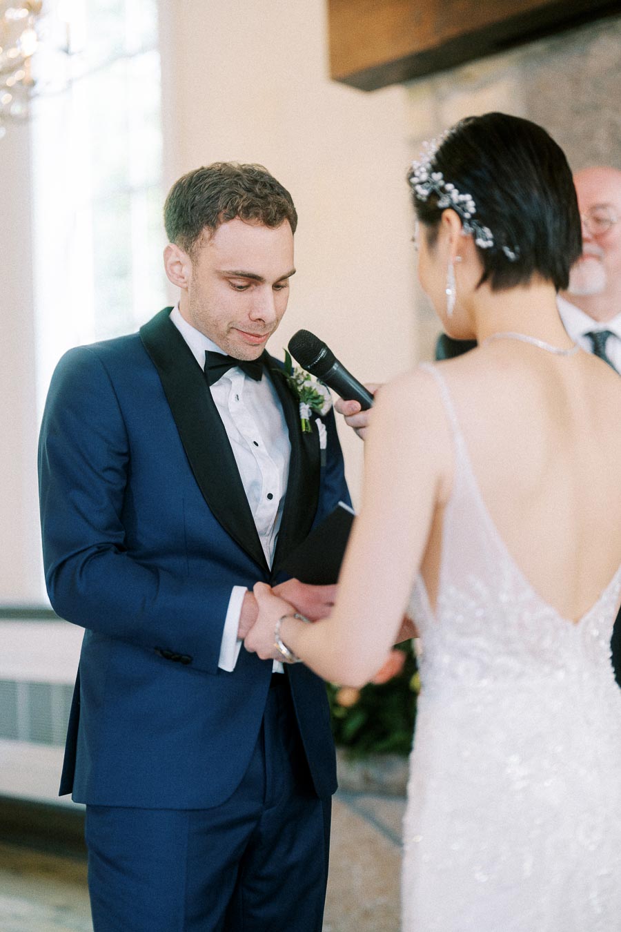 Groom in a blue tuxedo holds the bride’s hands as she reads vows into a microphone during their ceremony.