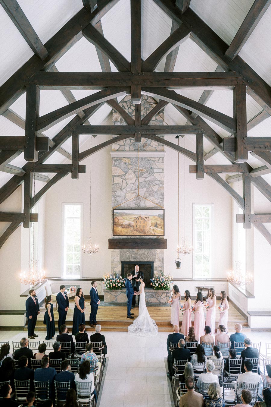Bride and groom stand at the altar during a chapel ceremony beneath exposed wooden beams and chandeliers.