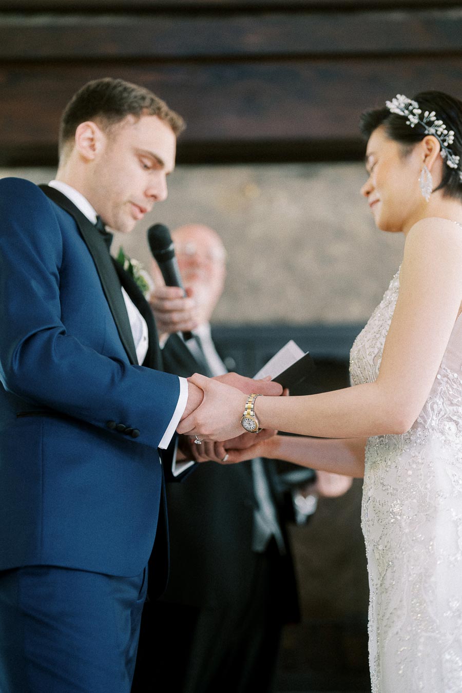 Groom in blue tuxedo holds the bride’s hands while reading vows during their indoor wedding ceremony.