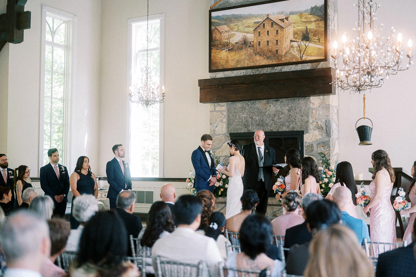 Bride and groom exchange rings during an indoor chapel ceremony before seated guests and attendants.