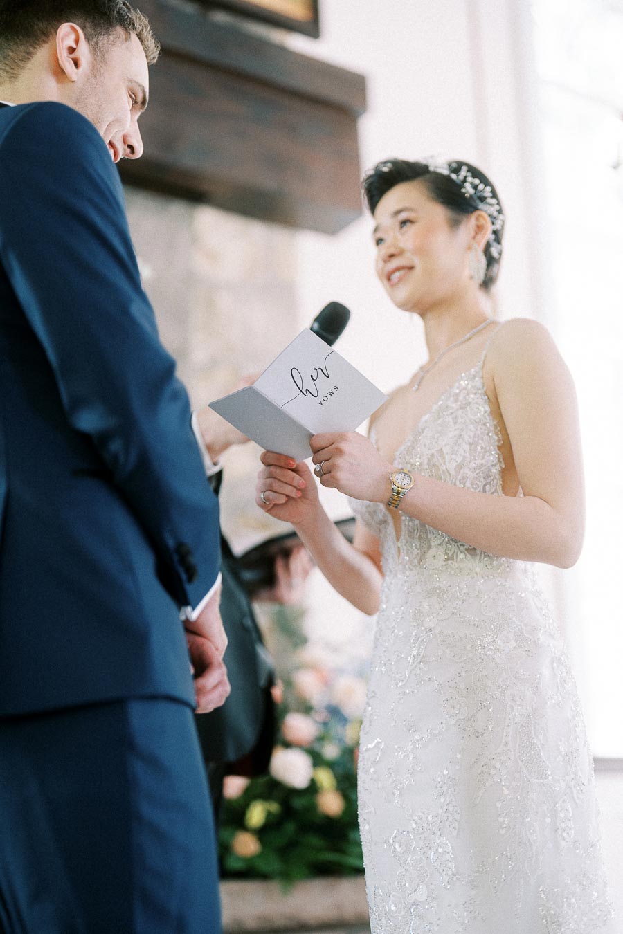 Bride reads vows from a booklet into a microphone while standing beside the groom during their ceremony.