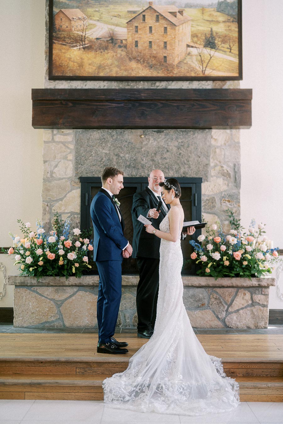 Bride reads vows to groom during an indoor ceremony before a stone fireplace with pastel floral arrangements.