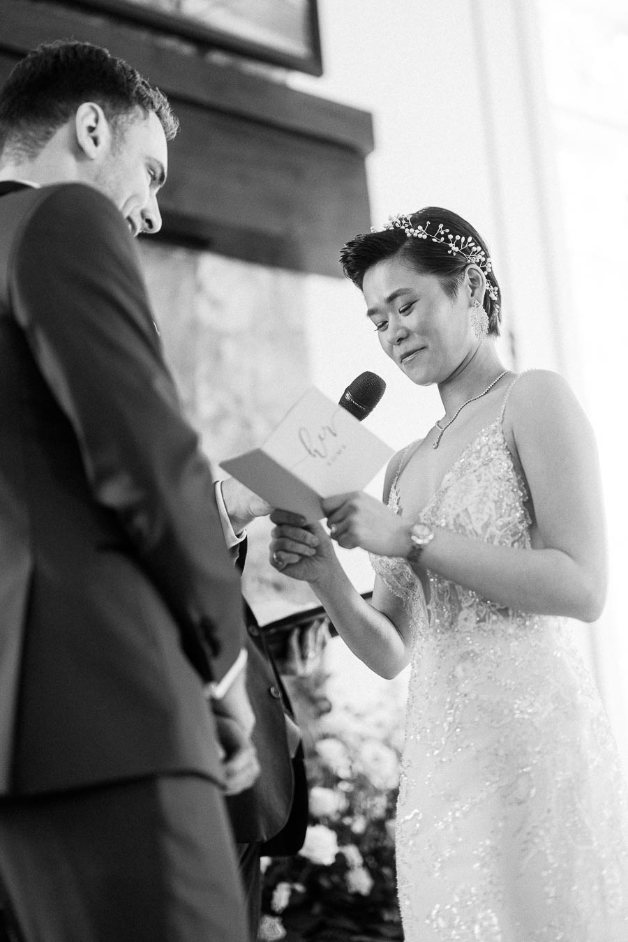 Miffy reads vows from a card into a microphone as Rawle smiles beside her during their wedding ceremony.