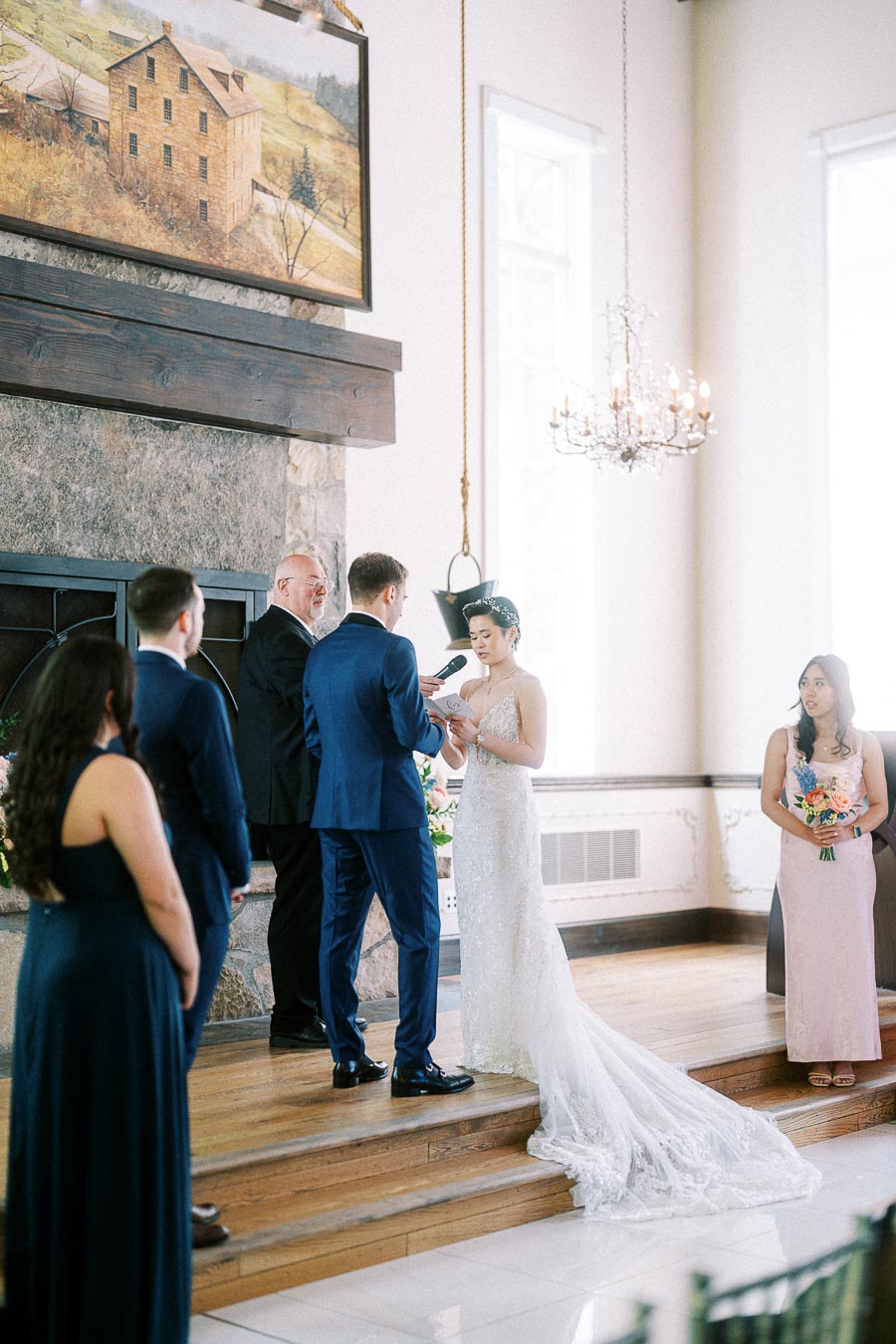 Bride and groom exchange rings beside an officiant and wedding party in a bright Ancaster Mill ceremony room.