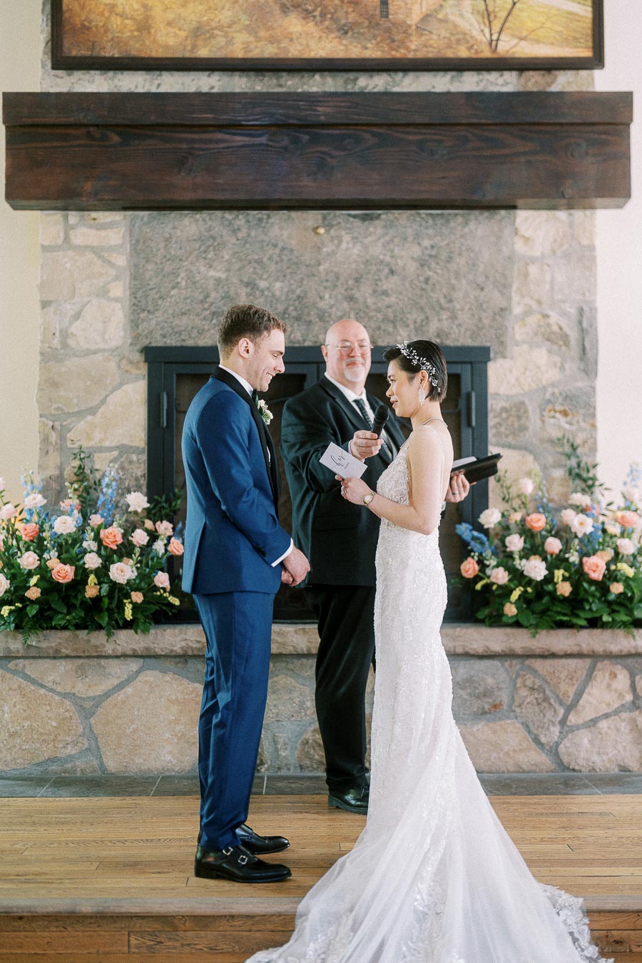 Miffy and Rawle exchange vows before an officiant in front of a stone fireplace with pastel floral arrangements.