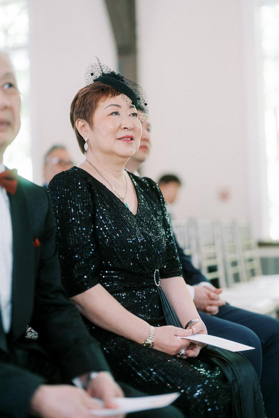 Woman in a black sequined dress and fascinator seated among guests during an indoor wedding ceremony