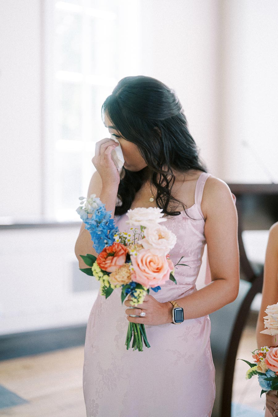 Bridesmaid in a pink dress wiping tears while holding a pastel bouquet beside a bright window