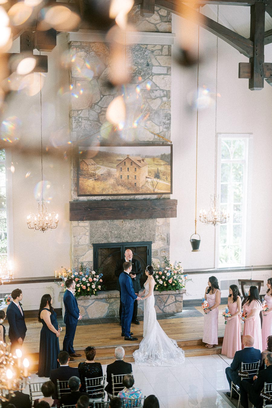 Miffy and Rawle stand at the altar during their indoor chapel ceremony beneath chandeliers and a stone fireplace.