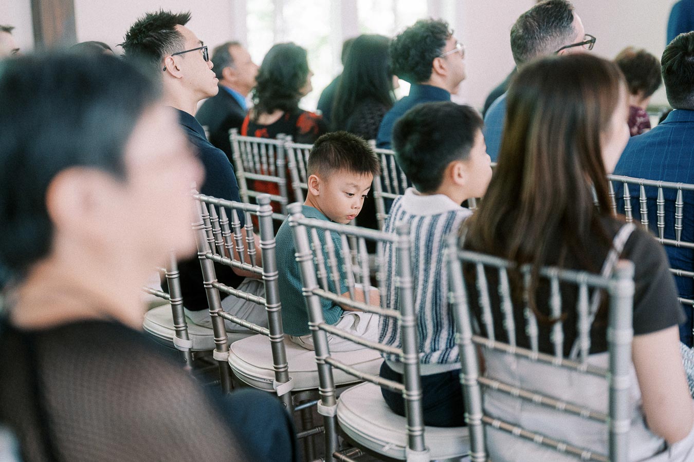 Wedding guests seated on silver chairs, with a young boy looking down during an indoor ceremony