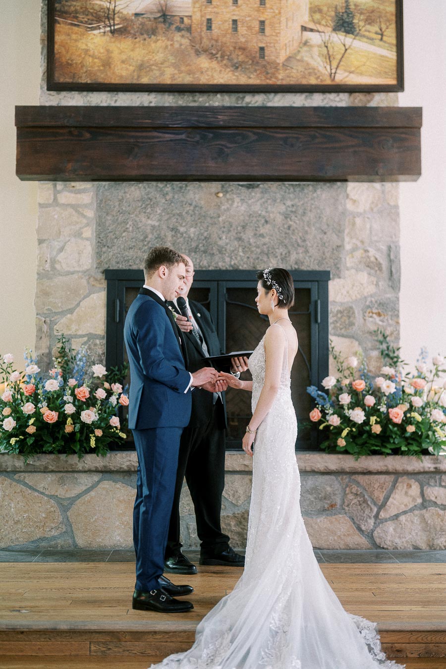 Bride and groom exchange rings during an indoor fireplace wedding ceremony with floral arrangements.