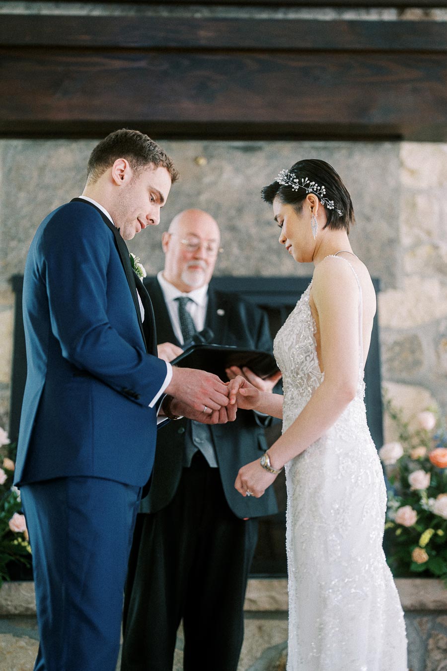 Groom places a wedding ring on the bride's finger during their indoor ceremony with an officiant behind them.
