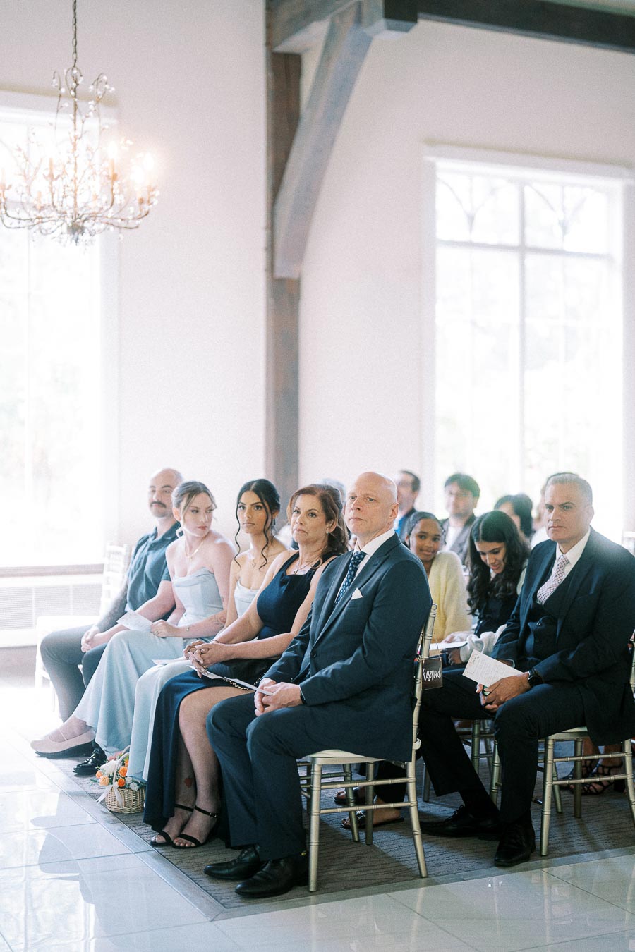 Wedding guests seated in a bright ceremony room with tall windows, chandelier, and gold chairs.
