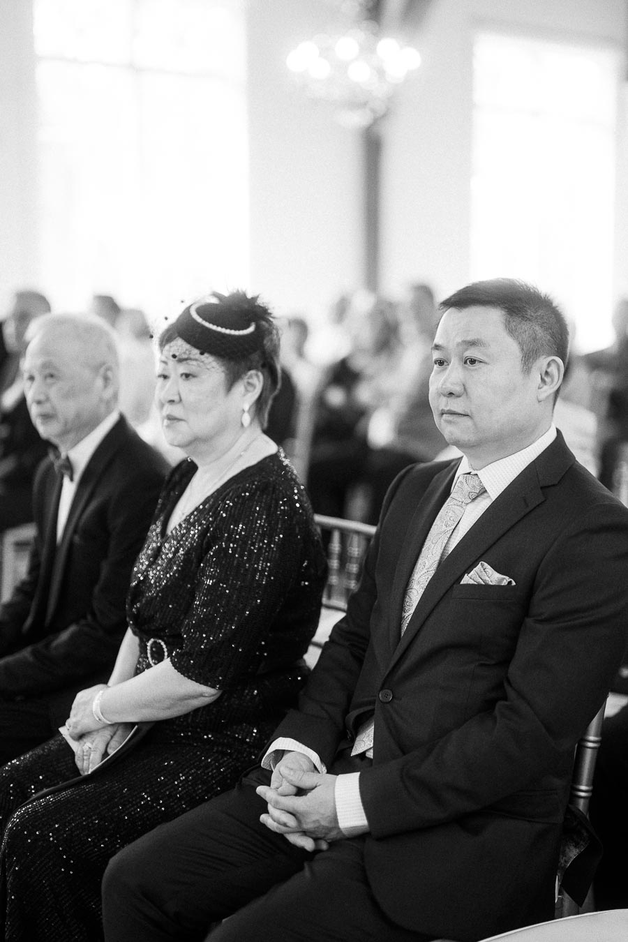 Three formally dressed wedding guests sit in a row during an indoor ceremony in black and white.