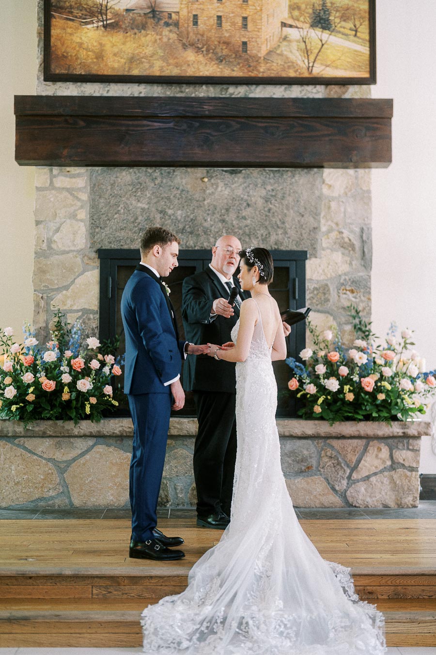 Miffy and Rawle hold hands during their indoor wedding ceremony before a stone fireplace with floral arrangements.