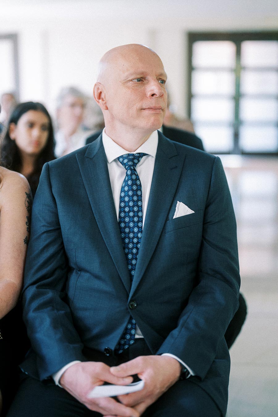 Seated wedding guest in a blue suit and patterned tie looks ahead during an indoor ceremony.