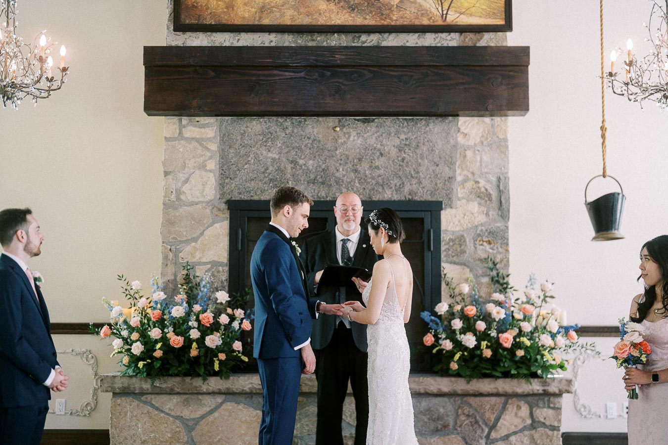 Bride and groom exchange rings before an officiant at a stone fireplace altar with floral arrangements.