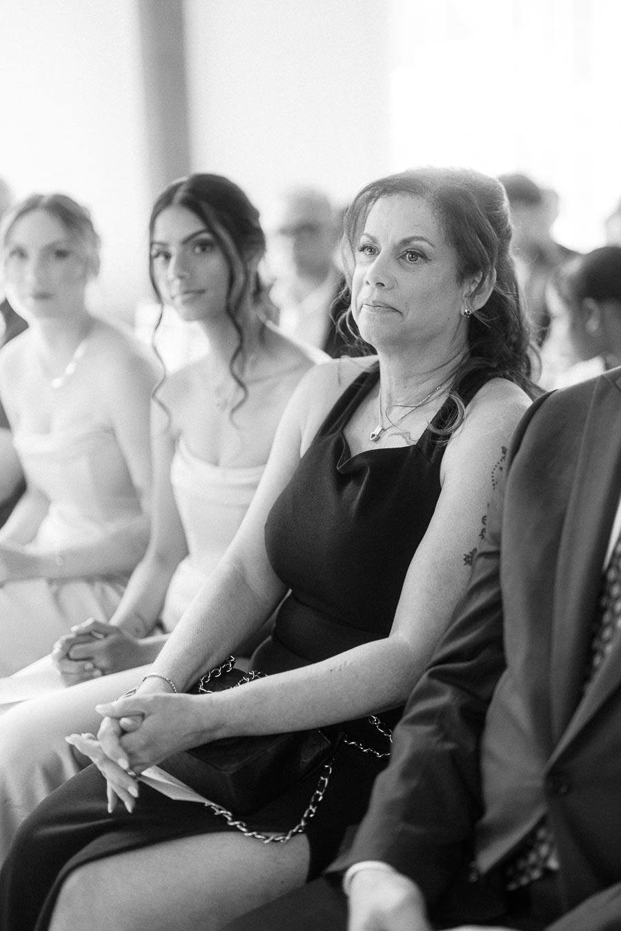 Black-and-white view of wedding guests seated indoors, including a woman in a black dress and bridesmaids nearby.